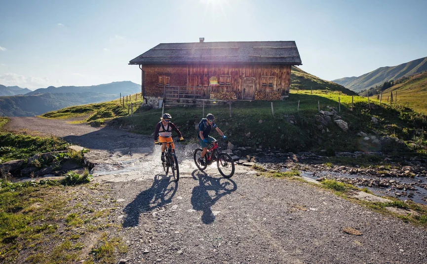 Zwei Menschen fahren mit Mountainbikes an einem Bach vor einer Holzhütte vorbei