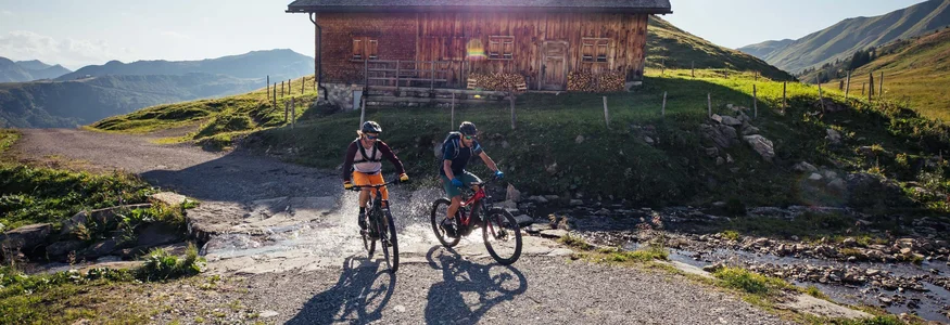 Zwei Menschen fahren mit Mountainbikes an einem Bach vor einer Holzhütte vorbei