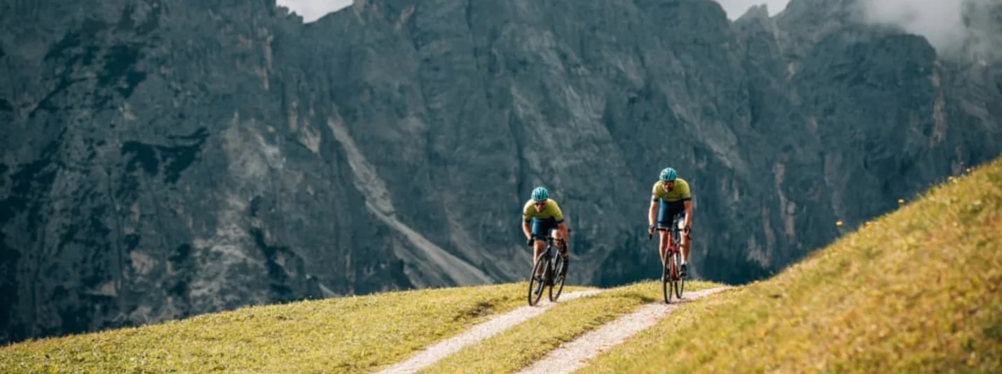 Two cyclists riding on a mountain trail with rocky cliffs in the background