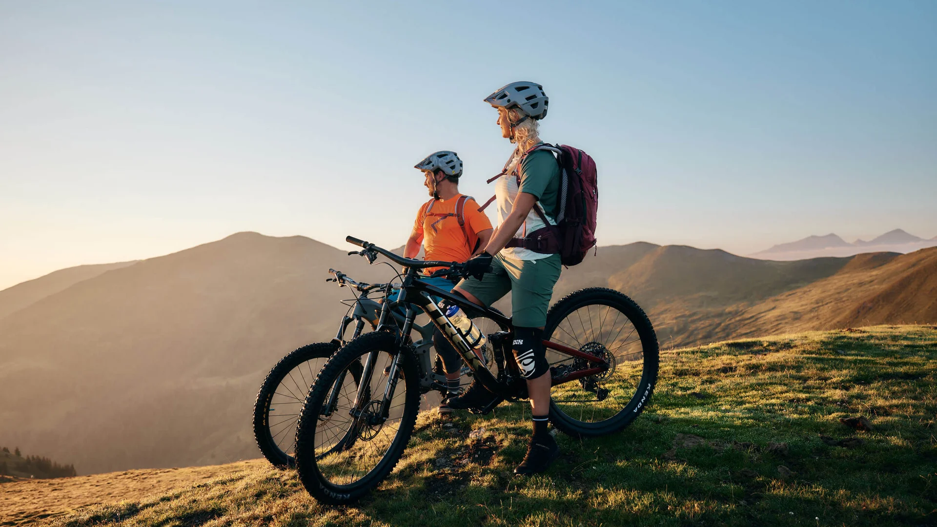 Two mountain bikers with helmets stand on a mountain with bikes at sunset