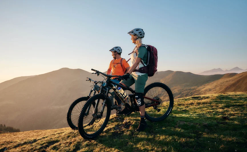 Two mountain bikers with helmets stand on a mountain with bikes at sunset