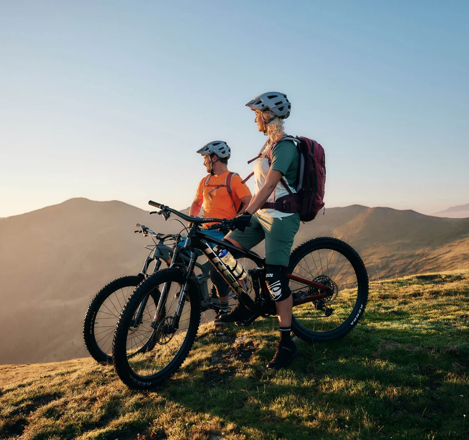 Zwei Mountainbiker mit Helmen stehen mit Fahrrädern auf einem Berg bei Sonnenuntergang