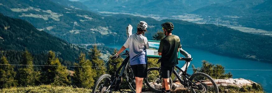 Twee fietsers lezen een informatiebord met uitzicht op bergen en meer