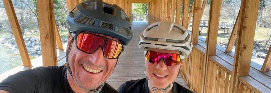 Bike und Hike – der alpine Trendsport, der das Beste aus zwei Welten vereint Zwei Radfahrer auf einer überdachten Holzbrücke lächelnd mit Helm und Sonnenbrille