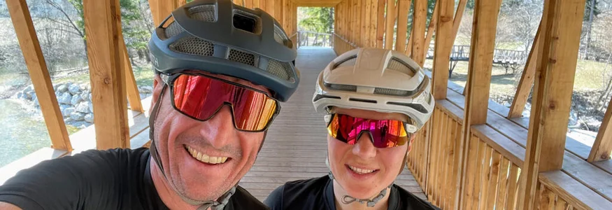 Zwei Radfahrer auf einer überdachten Holzbrücke lächelnd mit Helm und Sonnenbrille