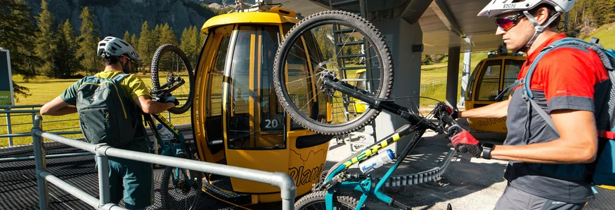 Two cyclists loading mountain bikes onto yellow cable car gondolas in the mountains