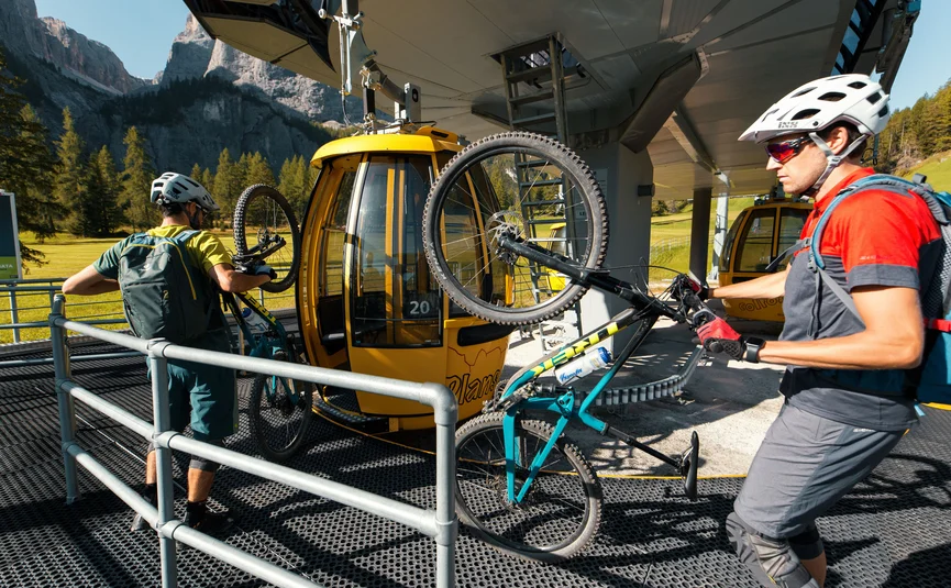 Two cyclists loading mountain bikes onto yellow cable car gondolas in the mountains