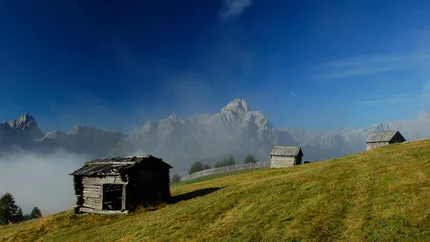 Almhütten auf grüner Wiese mit Bergen im Nebel unter blauem Himmel