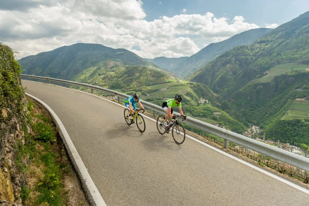Two cyclists riding on mountain road with green hills and cloudy sky