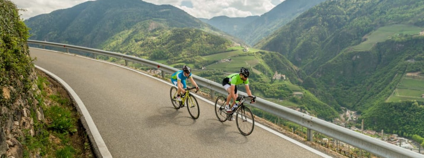 Two cyclists riding on mountain road with green hills and cloudy sky