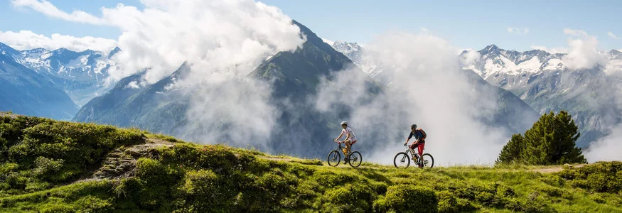 Zwei Radfahrer auf einem Bergweg mit Wolken und schneebedeckten Gipfeln im Hintergrund