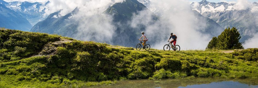 Zwei Radfahrer auf einem Bergweg mit Wolken und schneebedeckten Gipfeln im Hintergrund
