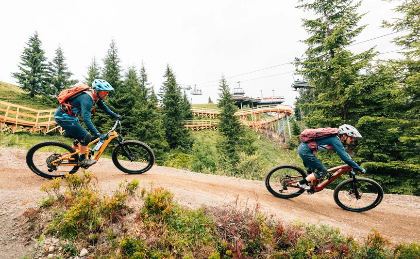 Zwei Mountainbiker auf einem Waldweg mit Bäumen und einer Seilbahn im Hintergrund