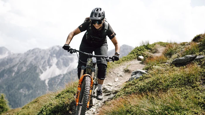 Frau fährt Mountainbike auf schmalem Gebirgspfad mit Bergen im Hintergrund