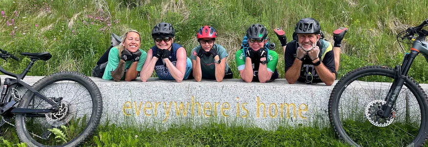 Fünf Radfahrer lächeln auf einer Bank mit der Aufschrift überall ist Heimat