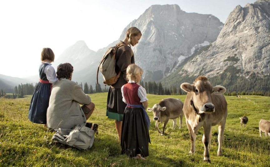 Family in traditional clothes watching cows on a green alpine meadow with mountains