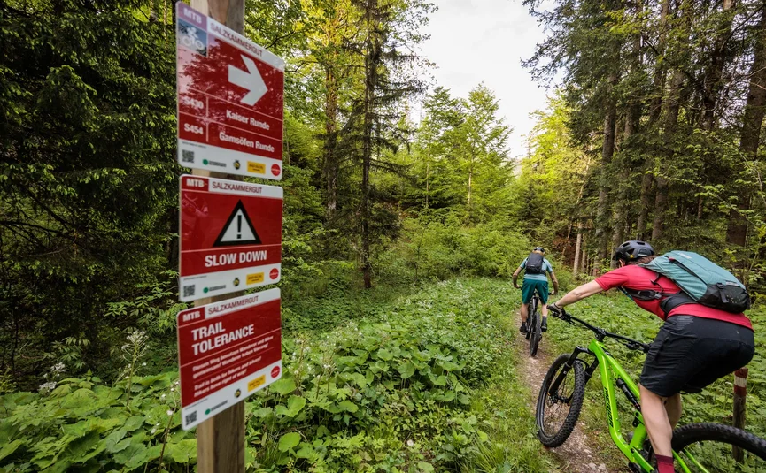 Zwei Mountainbiker auf Waldpfad neben Schildern mit Hinweisen und Warnungen
