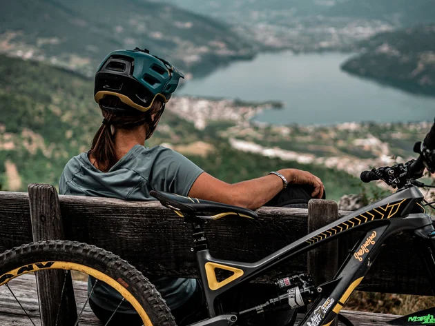 Frau mit Fahrradhelm sitzt auf Bank und blickt auf See und Berge