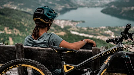 Frau mit Fahrradhelm sitzt auf Bank und blickt auf See und Berge