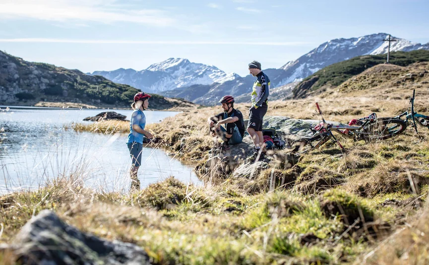 Drei Mountainbiker machen Pause an einem Bergsee mit verschneiten Bergen im Hintergrund