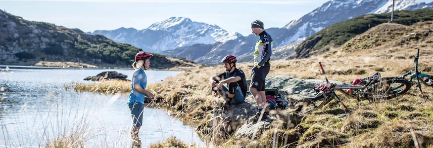Biken & Genießen im Montanara © Salzburger Sportwelt Drei Mountainbiker machen Pause an einem Bergsee mit verschneiten Bergen im Hintergrund