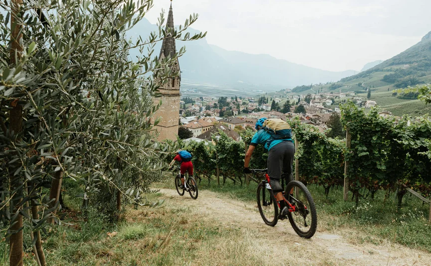 Two cyclists riding through a vineyard village with church and mountains in view