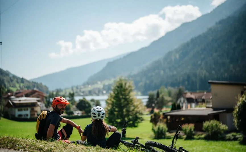 Zwei Radfahrer mit Helmen sitzen auf einer Wiese mit Berg- und Seeblick