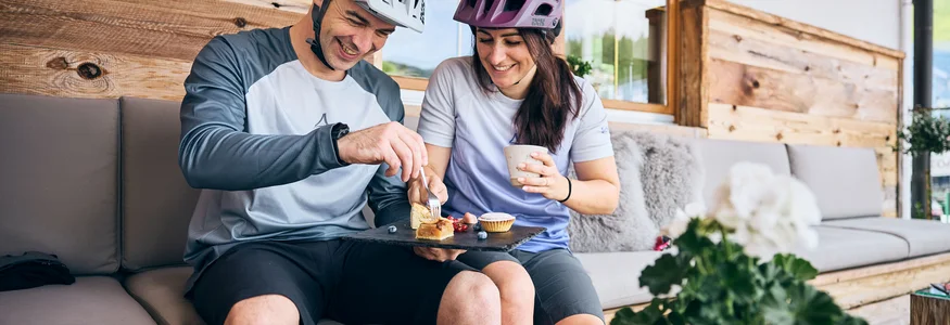 Bike-Region Flachau – von Radabenteuer bis Signature Ride © Tobias Köhler Paar mit Fahrradhelmen teilt Kuchen und Kaffee auf einer Terrasse