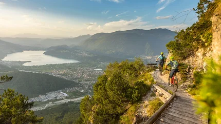 Two mountain bikers ride a trail overlooking a valley and lake