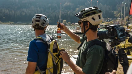 Zwei Radfahrer am See, einer hält eine Flasche Bier und öffnet einen Rucksack
