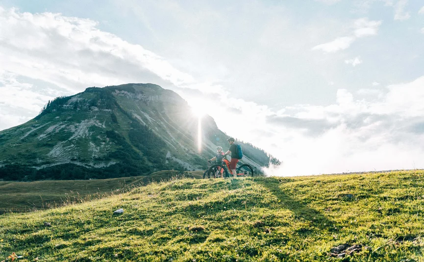 Twee fietsers staan op een groene heuvel met berg en zon op de achtergrond