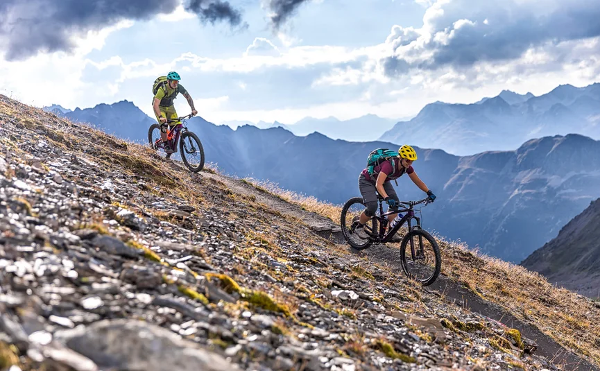 Zwei Mountainbiker fahren auf einem Bergpfad mit Alpen im Hintergrund