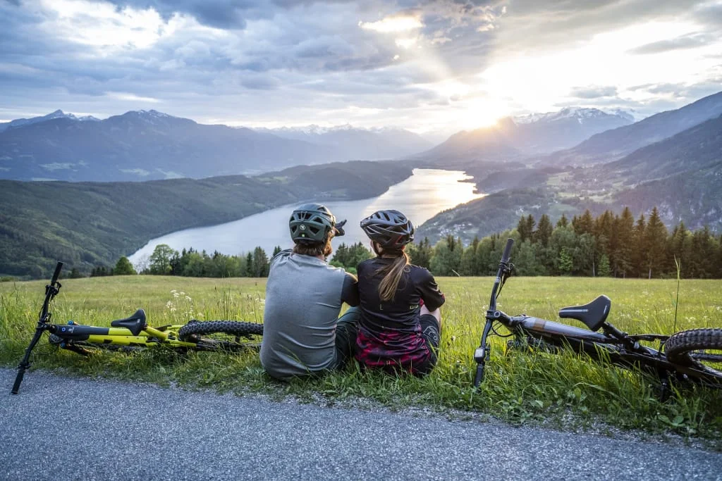 Zwei Radfahrer sitzen auf einer Wiese und betrachten einen See und Berge bei Sonnenuntergang
