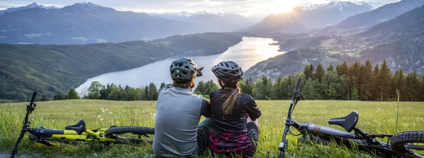 Twee fietsers zitten op gras en kijken naar een meer en bergen bij zonsondergang