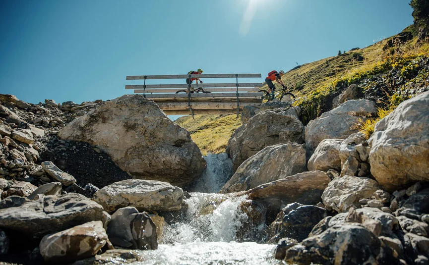 Zwei Mountainbiker fahren über eine Brücke über einen Bach in den Bergen