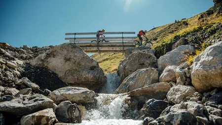Zwei Mountainbiker fahren über eine Brücke über einen Bach in den Bergen