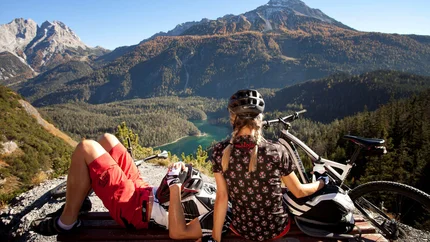 Zwei Mountainbiker auf einer Bank mit Blick auf Berge und See