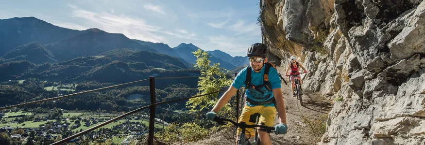 Twee mountainbikers op smal bergpad met zonnige hemel en uitzicht op dal