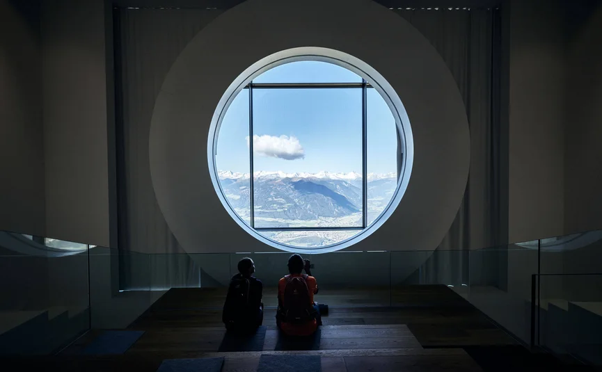 Zwei Personen sitzen vor einem großen runden Fenster mit Bergblick