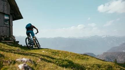 Radfahrer auf Bergpfad neben Holzhütte mit Bergpanorama