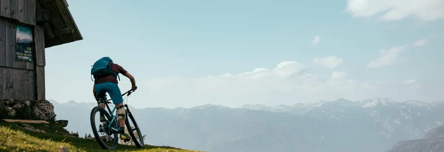 Radfahrer auf Bergpfad neben Holzhütte mit Bergpanorama