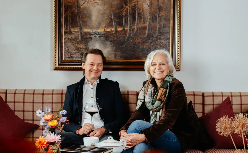 Zwei lächelnde Personen sitzen auf einem Sofa vor einem Waldgemälde