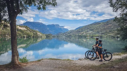 Two cyclists stand by lake looking at mountains and sky