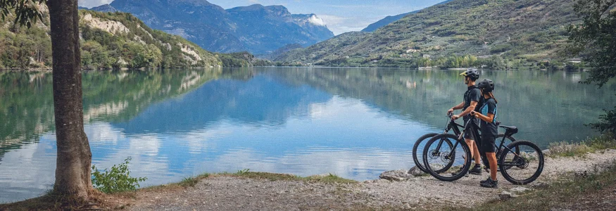 Two cyclists stand by lake looking at mountains and sky