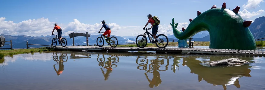 Drei Radfahrer auf einem Steg neben grünem Drachenskulptur am Bergsee