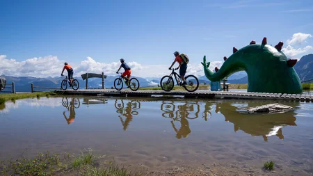 Ein Herbst-Wochenende in San Vigilio © Bike School San Vigilio - Dragon Trail am Kronplatz mit den Guides der Bike School San Vigilio Drei Radfahrer auf einem Steg neben grünem Drachenskulptur am Bergsee
