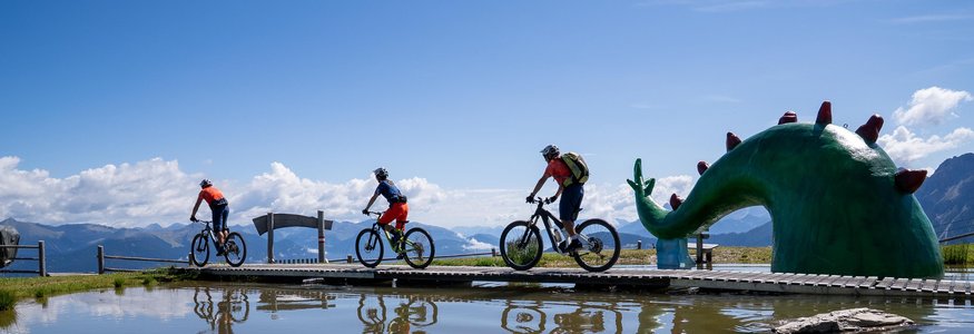 Drei Radfahrer auf einem Steg neben grünem Drachenskulptur am Bergsee