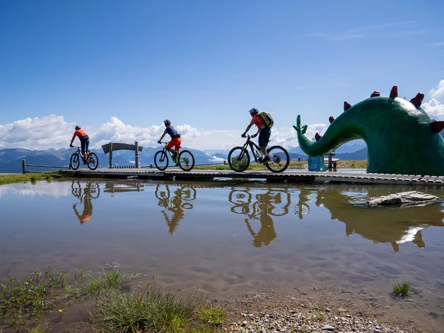 Ein Herbst-Wochenende in San Vigilio © Bike School San Vigilio - Dragon Trail am Kronplatz mit den Guides der Bike School San Vigilio Drei Radfahrer auf einem Steg neben grünem Drachenskulptur am Bergsee
