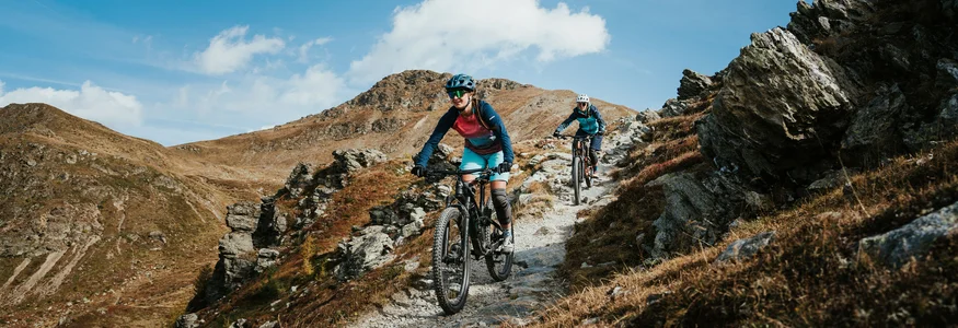 Zwei Mountainbiker fahren auf einem felsigen Bergweg unter blauem Himmel