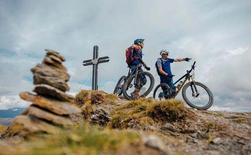 Zwei Fahrradfahrer mit Helmen auf einem Berggipfel neben einem Kreuz